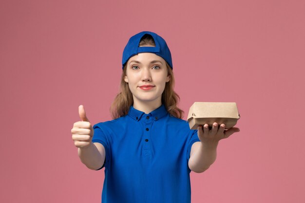 Corriere femminile di vista frontale in uniforme blu e mantello che tiene piccolo pacchetto di cibo di consegna sulla parete rosa, società di servizi uniformi di consegna di lavoro