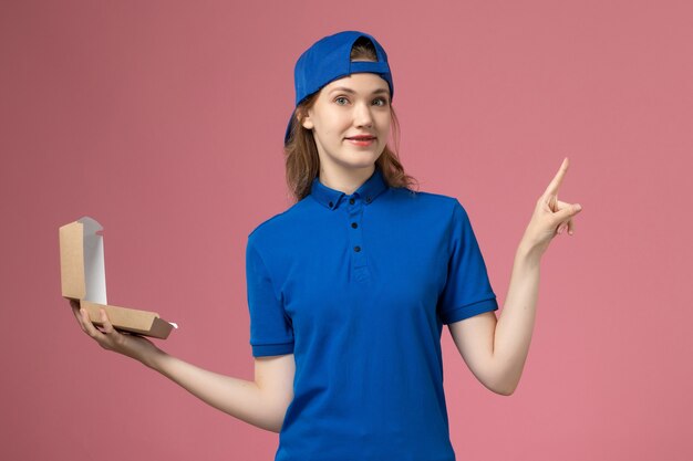 Corriere femminile di vista frontale in uniforme blu e mantello che tiene poco pacchetto di cibo di consegna sulla parete rosa chiaro, lavoratore dipendente di servizio uniforme di consegna