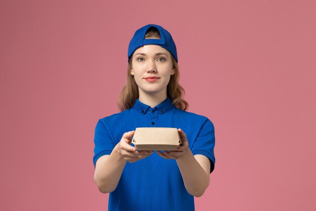 Corriere femminile di vista frontale in uniforme blu e mantello che tiene poco pacchetto di cibo di consegna sullo sfondo rosa uniforme di consegna servizio azienda lavoro lavoratore ragazza lavoro