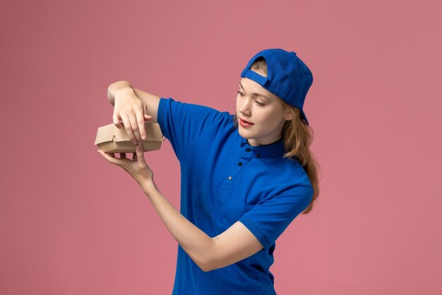 Corriere femminile di vista frontale in uniforme blu e mantello che tiene un piccolo pacchetto di cibo per la consegna e aprendolo sul muro rosa, società di servizi di consegna uniforme
