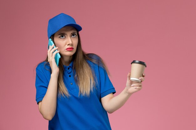 Corriere femminile di vista frontale in uniforme blu e tazza di caffè di consegna della tenuta del capo che parla sul telefono sulla parete rosa