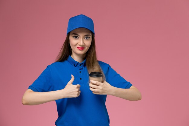 Corriere femminile di vista frontale in uniforme blu e tazza di caffè di consegna della tenuta del capo che posa sulla parete rosa