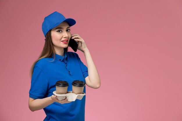 Corriere femminile di vista frontale in uniforme blu e tazze di caffè di consegna della tenuta del capo che parlano sul telefono sulla parete rosa