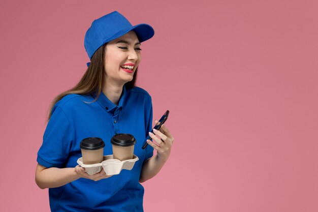 Corriere femminile di vista frontale in uniforme blu e tazze di caffè di consegna della tenuta del capo che ridono sulla parete rosa