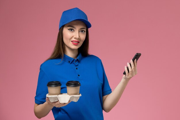 Corriere femminile di vista frontale in uniforme blu e tazze di caffè di consegna della tenuta del capo usando il suo telefono sulla parete rosa