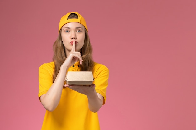 Corriere femminile di vista frontale in uniforme gialla e capo che tiene il pacchetto dell'alimento di consegna sul lavoro della ragazza dell'operaio dell'azienda dell'uniforme di consegna di servizio della parete rosa