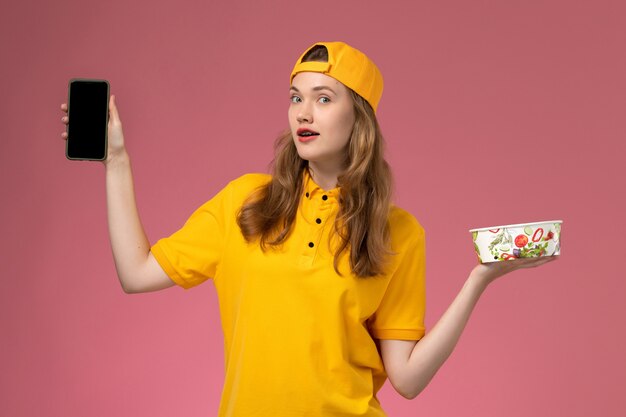 Corriere femminile di vista frontale in uniforme gialla e ciotola di consegna della tenuta del capo e telefono sul lavoratore dell'uniforme di consegna di servizio dell'azienda della parete rosa