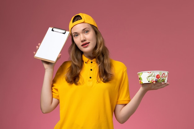 Corriere femminile di vista frontale in uniforme gialla e mantello che tiene ciotola di consegna e blocco note sul lavoratore dell'azienda uniforme di consegna servizio parete rosa chiaro