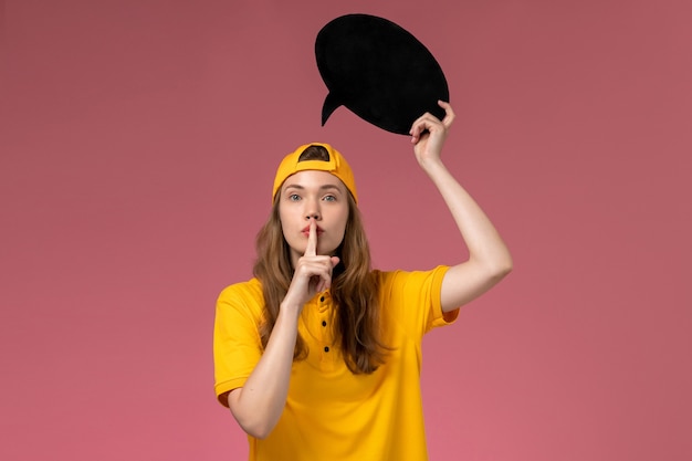 Corriere femminile di vista frontale in uniforme gialla e mantello che tiene grande segno nero sulla parete rosa, lavoro uniforme di consegna di lavoro di servizio dell'azienda