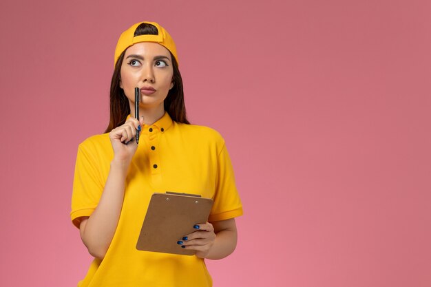 Corriere femminile di vista frontale in uniforme gialla e mantello che tiene il blocco note e la penna che pensano sul lavoro di consegna dell'uniforme di servizio dell'azienda della parete rosa chiaro