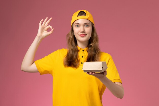 Corriere femminile di vista frontale in uniforme gialla e mantello che tiene il pacchetto di cibo di consegna sul lavoratore dell'azienda uniforme di consegna di servizio della parete rosa