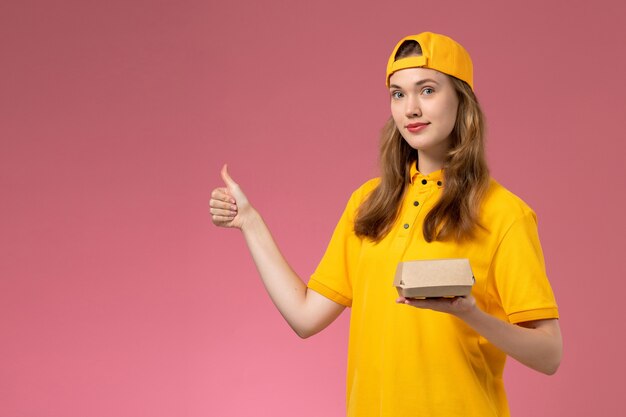 Corriere femminile di vista frontale in uniforme gialla e mantello che tiene il pacchetto di cibo di consegna sul lavoro dell'operaio dell'azienda dell'uniforme di consegna di servizio della parete rosa