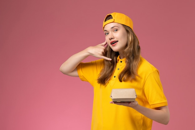 Corriere femminile di vista frontale in uniforme gialla e mantello che tiene il pacchetto di cibo di consegna sul lavoro della ragazza dell'operaio dell'uniforme di consegna di servizio della parete rosa