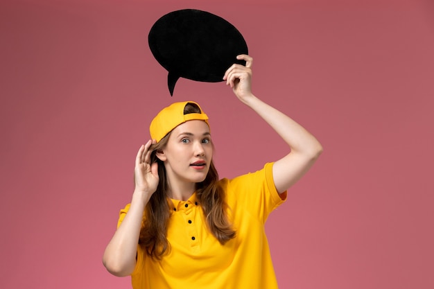 Corriere femminile di vista frontale in uniforme gialla e mantello che tiene il segno nero sul lavoratore della ragazza di lavoro dell'uniforme di consegna di servizio dell'azienda della parete rosa