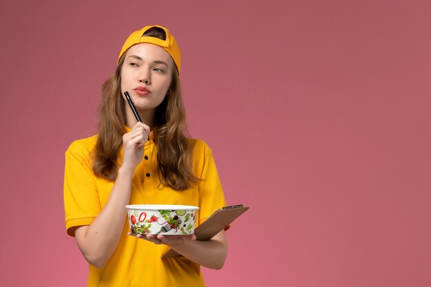 Corriere femminile di vista frontale in uniforme gialla e mantello che tiene la ciotola di consegna e il blocco note pensando sulla società di lavoro uniforme di consegna del lavoratore di servizio della parete rosa