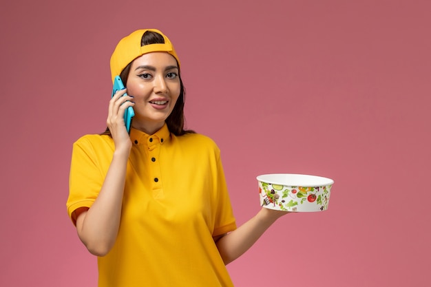 Corriere femminile di vista frontale in uniforme gialla e mantello che tiene la ciotola di consegna e utilizzando un telefono sul lavoratore di consegna uniforme di servizio parete rosa chiaro