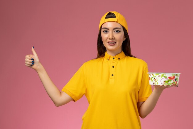 Corriere femminile di vista frontale in uniforme gialla e mantello che tiene la ciotola di consegna rotonda sul lavoro di consegna dell'uniforme di servizio della parete rosa