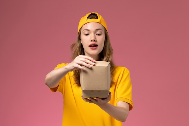 Corriere femminile di vista frontale in uniforme gialla e mantello che tiene piccoli pacchetti di cibo di consegna sull'uniforme di consegna di servizio della parete rosa chiaro