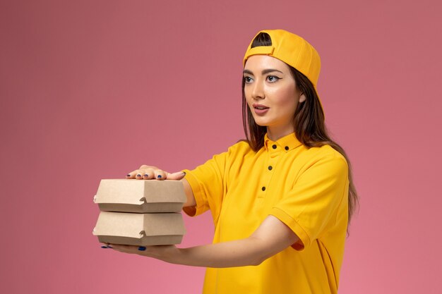 Corriere femminile di vista frontale in uniforme gialla e mantello che tiene piccoli pacchi di cibo di consegna sul lavoro della ragazza di consegna uniforme di servizio della parete rosa chiaro