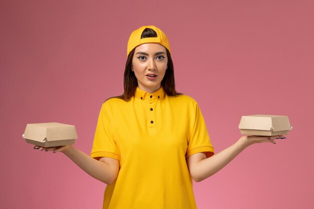 Corriere femminile di vista frontale in uniforme gialla e mantello che tiene piccoli pacchi di cibo di consegna sul lavoro di consegna uniforme di servizio da parete rosa chiaro