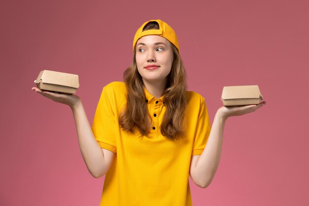 Corriere femminile di vista frontale in uniforme gialla e mantello che tiene piccoli pacchi di cibo per la consegna su uniforme di consegna di lavoro di servizio di parete rosa chiaro