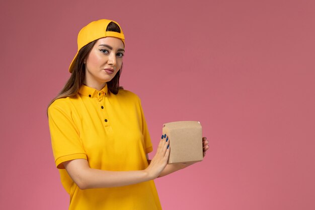 Corriere femminile di vista frontale in uniforme gialla e mantello che tiene piccolo pacchetto dell'alimento di consegna sulla società di consegna del lavoratore di servizio dell'uniforme della parete rosa chiaro