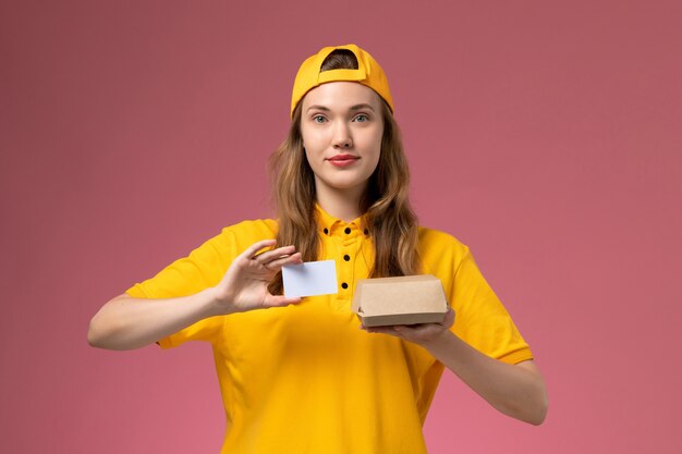 Corriere femminile di vista frontale in uniforme gialla e mantello che tiene piccolo pacchetto di cibo di consegna e carta di plastica sul lavoratore uniforme di consegna di servizio di lavoro della parete rosa