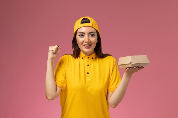 Corriere femminile di vista frontale in uniforme gialla e mantello che tiene piccolo pacchetto di cibo di consegna sul lavoro dell'azienda di consegna di servizio uniforme della parete rosa