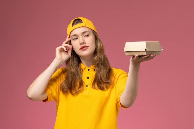 Corriere femminile di vista frontale in uniforme gialla e mantello che tiene piccolo pacchetto di cibo di consegna sul lavoro uniforme di consegna di servizio della parete rosa