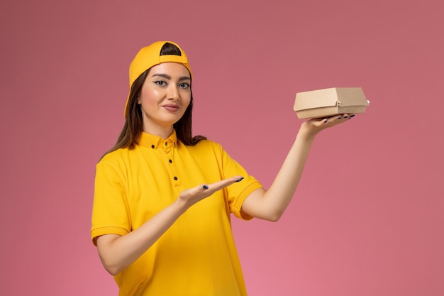 Corriere femminile di vista frontale in uniforme gialla e mantello che tiene piccolo pacchetto di cibo di consegna sulla ragazza della società di consegna di servizio uniforme della parete rosa