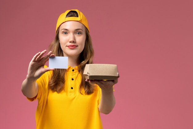Corriere femminile di vista frontale in uniforme gialla e mantello che tiene piccolo pacchetto di cibo per la consegna e carta di plastica sull'uniforme di consegna del servizio di parete rosa