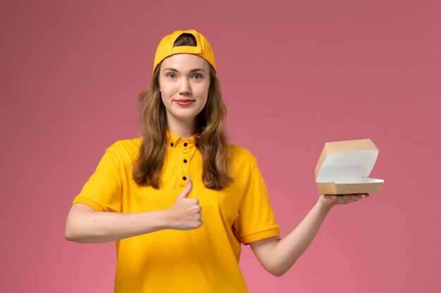 Corriere femminile di vista frontale in uniforme gialla e mantello che tiene piccolo pacchetto di cibo vuoto di consegna sull'uniforme di consegna del servizio di parete rosa chiaro