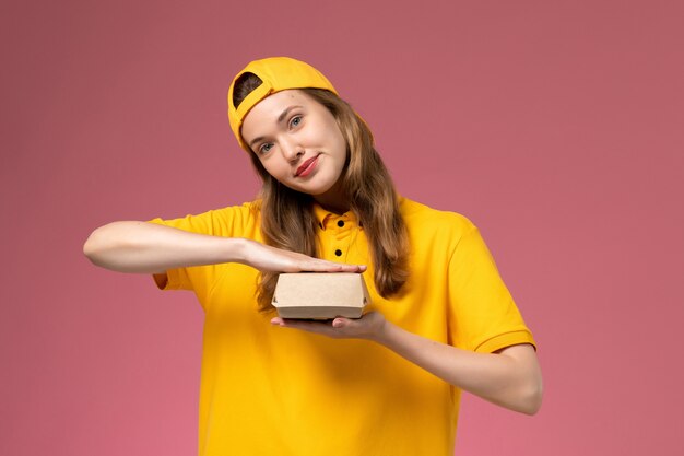 Corriere femminile di vista frontale in uniforme gialla e mantello che tiene piccolo pacchetto di cibo vuoto di consegna sull'uniforme di consegna del servizio di parete rosa chiaro