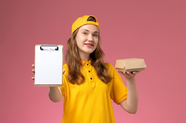 Corriere femminile di vista frontale in uniforme gialla e mantello che tiene poco pacchetto di cibo di consegna e blocco note sul lavoro uniforme di consegna di servizio di parete rosa chiaro