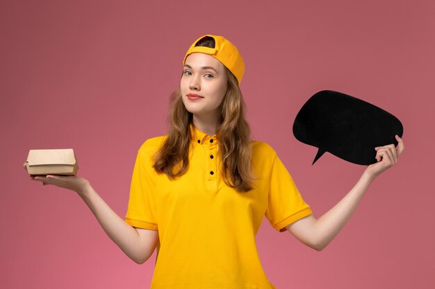 Corriere femminile di vista frontale in uniforme gialla e mantello che tiene segno nero con il pacchetto di cibo sull'uniforme di consegna di servizio dell'azienda della parete rosa