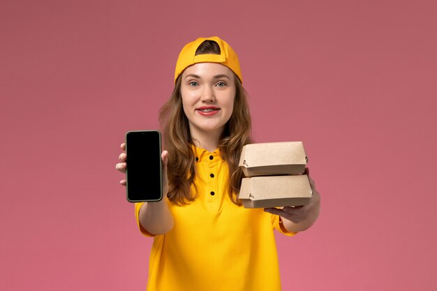 Corriere femminile di vista frontale in uniforme gialla e telefono della tenuta del capo con i pacchetti dell'alimento sul lavoro dell'uniforme di consegna di servizio dell'azienda della parete rosa