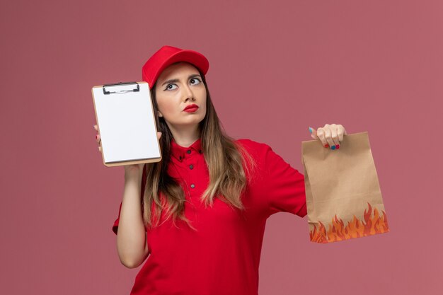 Corriere femminile di vista frontale in uniforme rossa che tiene blocco note e pacchetto di cibo pensando sulla società uniforme di consegna del lavoro di servizio sfondo rosa