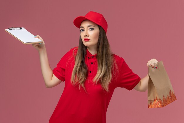 Corriere femminile di vista frontale in uniforme rossa che tiene blocco note e pacchetto di cibo su sfondo rosa azienda uniforme di lavoro di consegna servizio