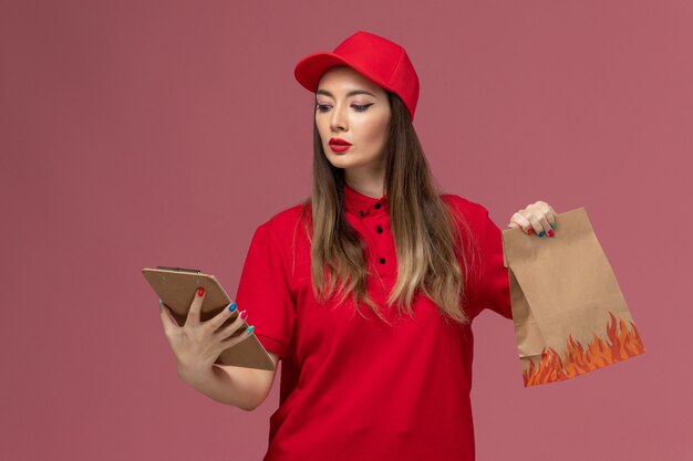 Corriere femminile di vista frontale in uniforme rossa che tiene blocco note e pacchetto di cibo sulla società uniforme di consegna di lavoro servizio scrivania rosa