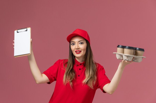 Corriere femminile di vista frontale in uniforme rossa che tiene il blocco note delle tazze di caffè di consegna che sorride sull'uniforme di lavoro della società di consegna del servizio del fondo rosa