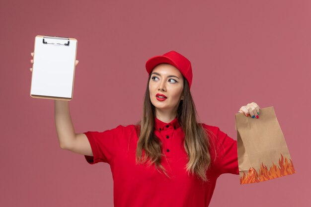 Corriere femminile di vista frontale in uniforme rossa che tiene il blocco note e il pacchetto di cibo sulla società uniforme di consegna del lavoro di servizio di sfondo rosa