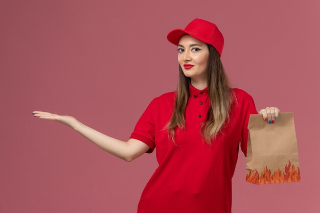 Corriere femminile di vista frontale in uniforme rossa che tiene il pacchetto alimentare di carta che sorride sull'azienda dell'uniforme di consegna di lavoro di servizio del fondo rosa