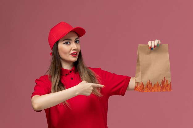 Corriere femminile di vista frontale in uniforme rossa che tiene il pacchetto alimentare di carta sulla società uniforme di consegna del lavoro di servizio sfondo rosa chiaro