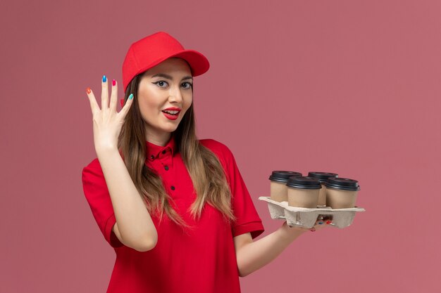 Corriere femminile di vista frontale in uniforme rossa che tiene le tazze di caffè di consegna che mostrano le sue unghie sul lavoratore uniforme di lavoro di consegna del servizio di sfondo rosa