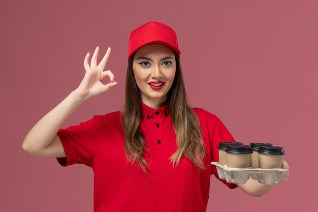 Corriere femminile di vista frontale in uniforme rossa che tiene le tazze di caffè di consegna e che sorride sull'operaio di lavoro uniforme di consegna di servizio del fondo rosa