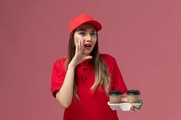 Corriere femminile di vista frontale in uniforme rossa che tiene le tazze di caffè di consegna marroni che bisbigliano su sfondo rosa chiaro servizio di consegna uniforme lavoratore lavoro azienda femmina