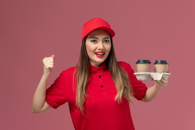 Corriere femminile di vista frontale in uniforme rossa che tiene le tazze di caffè di consegna sullo sfondo rosa uniforme di consegna del servizio di lavoro del lavoratore