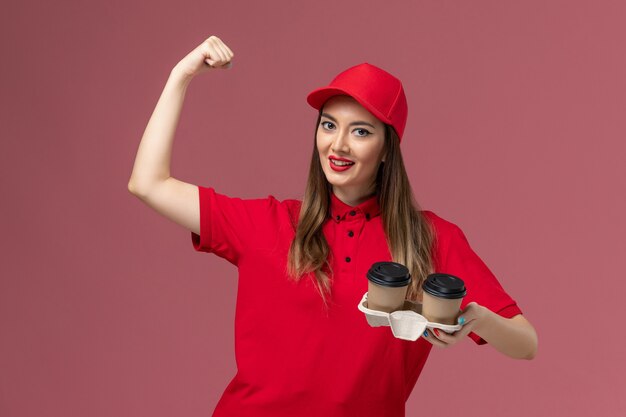 Corriere femminile di vista frontale in uniforme rossa che tiene le tazze di caffè marroni di consegna che flettono sull'azienda dell'uniforme di consegna di servizio del fondo rosa chiaro