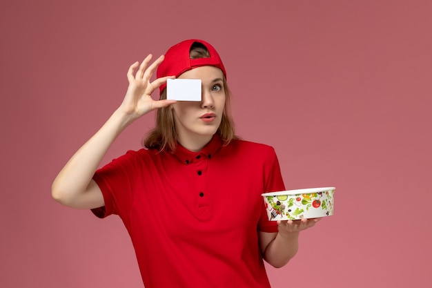 Corriere femminile di vista frontale in uniforme rossa e ciotola di consegna della tenuta del capo con la carta sulla parete rosa-chiaro, consegna dell'uniforme del lavoro di servizio del lavoratore