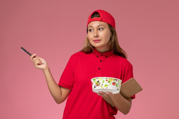 Corriere femminile di vista frontale in uniforme rossa e mantello che tiene blocco note e ciotola di consegna sul servizio di consegna uniforme di lavoro sfondo rosa